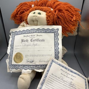 May include: A vintage Cabbage Patch Kid doll with orange yarn hair and a cream-colored face. The doll holds a birth certificate and adoption papers, both with the name "Abigail Sophia" and the date "September 17, 1981".