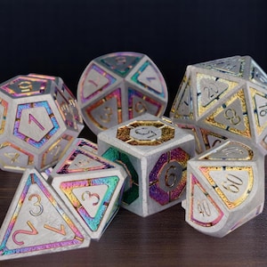 Puede incluir: Un conjunto de siete dados de metal iridiscente con números dorados y blancos. Los dados son de varias formas y tamaños, incluyendo un d4, d6, d8, d10, d12 y d20.