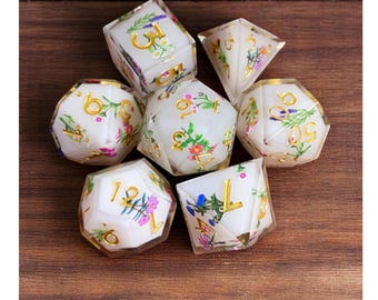 May include: Set of seven polyhedral dice with a floral design. The dice are white with gold numbers and colorful flowers. The dice are arranged on a wooden surface. The dice are for tabletop games.