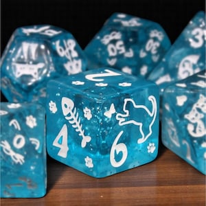 Könnte beinhalten: Ein Satz von sieben blauen Harzwürfeln mit weißen Zahlen und Katzenmotiven. Die Würfel zeigen Katzensilhouetten, Pfotenabdrücke und Fischgräten. Die Würfel werden auf einer Holzoberfläche präsentiert.