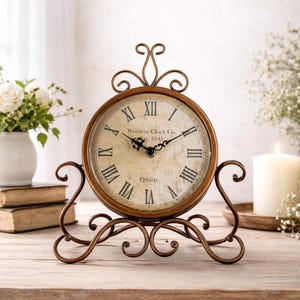 Puede incluir: Un reloj de cuarzo de color bronce con números romanos y el texto "Winston Clock Co. Est. 1845." El reloj tiene una base y una parte superior decorativas con volutas. La esfera del reloj es blanquecina con agujas negras.