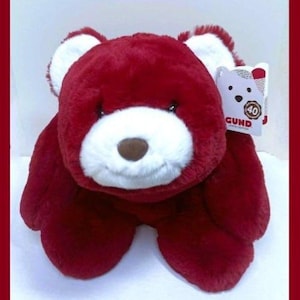 May include: A plush, red teddy bear with white accents on the ears and muzzle. The bear has a brown nose and a tag that reads "GUND". The bear is soft and cuddly, perfect for a child's toy collection.