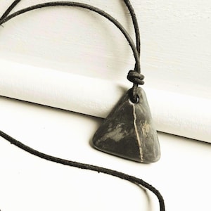Puede incluir: Collar con colgante triangular gris oscuro y cordón negro. El colgante tiene una franja de color claro y un agujero en la parte superior. El cordón está anudado por encima del colgante.