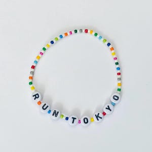 May include: A beaded bracelet with the words "RUN TOKYO" spelled out in white letter beads. The bracelet features a rainbow of colorful beads between the white letter beads. The bracelet is on a white background.