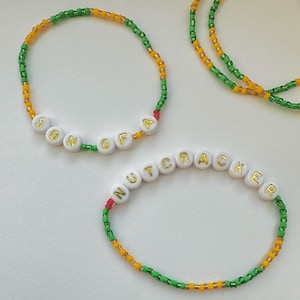 May include: Two beaded bracelets with green, yellow, and red beads. One bracelet spells "SON OF A" and the other spells "NUTCRACKER".