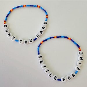 May include: Two beaded bracelets with white letter beads spelling "DON'T YOU DARE" and "SETTLE FOR FINE". The bracelets have blue, red, and yellow beads. The bracelets are on a white surface.