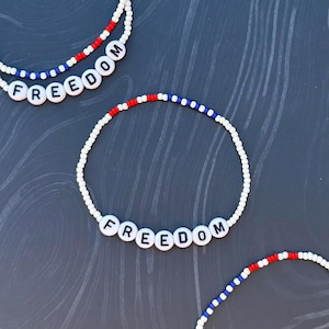 Könnte beinhalten: Weiße Perlenarmbänder mit roten, weißen und blauen Akzenten. Das Wort "FREEDOM" ist mit schwarzen Buchstabenperlen geschrieben. Die Armbänder liegen auf einer dunkelgrauen Oberfläche.