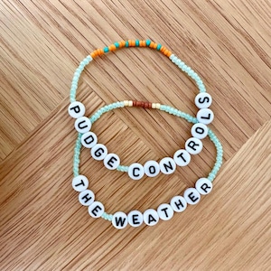 May include: Two beaded bracelets with the words "Pudge Controls The Weather" on them. The bracelets are made with light blue and orange beads.
