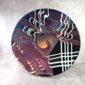 May include: A decorative ceramic bowl with a unique design. The bowl features a swirl pattern in the center, with shades of purple, blue, and gold. White and black accents create a striking contrast. The bowl is displayed on a clear stand.