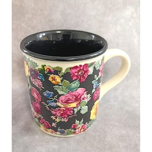 May include: A ceramic mug with a black interior and a cream-colored handle. The exterior is decorated with a floral pattern featuring pink, yellow, and purple flowers on a black background. The rim is black.