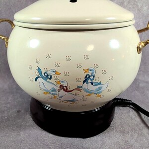 May include: A cream-colored electric pot with a matching lid and gold-tone metal handles. The exterior features a repeating pattern of small flowers and a central graphic of three white geese wearing blue and red ribbons. It sits on a black base.
