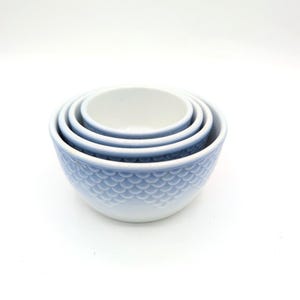 May include: A set of three nested ceramic bowls in a light blue color. Each bowl features a raised, repeating scale-like texture on the exterior surface. The interior of each bowl is smooth and white, with a light blue rim at the top edge.