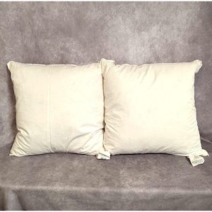 May include: Two off-white square pillows. The pillows are made of a soft, textured fabric. The pillows are placed on a gray surface. The pillows are a neutral color and would complement any bedroom decor.