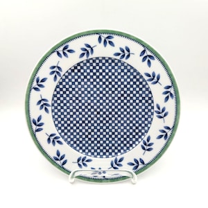 May include: A round ceramic plate featuring a central blue and white checkered pattern. The rim is decorated with a repeating blue leaf motif on a white background, framed by a thin green border along the outer edge of the dinnerware piece.
