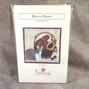 May include: A Harvest Bunny wool applique pattern in a clear plastic sleeve. The white card features the text Harvest Bunny, Finished block 8" x 8", and Wool Applique. The illustration shows a grey rabbit, a brown pumpkin, and a sunflower.