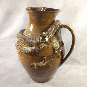May include: A brown ceramic pitcher with a sculpted snake wrapped around the body and handle. The snake has detailed scales and a small, open mouth. The pitcher has a wide base and a narrow neck.