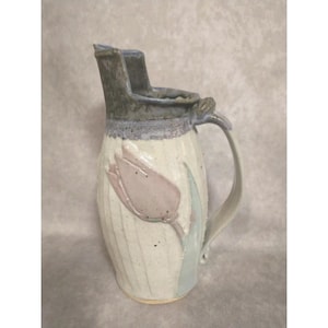 May include: A handmade ceramic pitcher with a tulip design. The pitcher is off-white with a textured surface and a light purple tulip. The handle and spout are gray-green. The pitcher is approximately 8 inches tall.