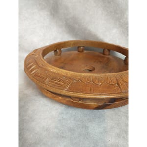 May include: A round, wooden decorative bowl with a carved floral design around the rim. The bowl is a warm brown color and has a raised, circular edge with small, rounded supports. The center of the bowl has a small hole.