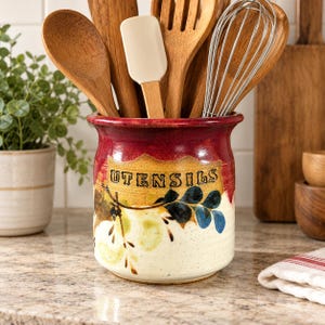 May include: A ceramic utensil crock with a red rim and a cream-colored base featuring a hand-painted botanical design and the word UTENSILS embossed in black. The crock is filled with various wooden spoons, a spatula, and a metal whisk.