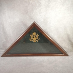 May include: Triangular wooden flag display case with a dark finish. The case features a glass front and a gold-colored emblem of an eagle with a shield and olive branches. The interior has a dark green fabric backing.