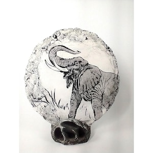 May include: A circular stone plaque featuring a black and white illustration of an elephant with its trunk raised. The plaque has a textured, cracked border and is supported by a dark, porous, rock-like base, creating a sculptural display piece.