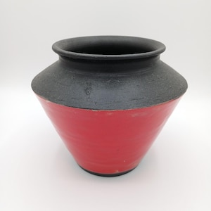 May include: A handcrafted ceramic vase with a striking two-tone design. The upper portion is a matte black, contrasting with the vibrant red lower section. The vase has a wide, rounded body and a slightly flared rim, suitable for home decor.