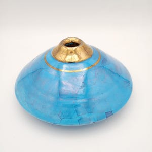 May include: A low-profile, wide-bodied vase featuring a vibrant turquoise blue mosaic tile exterior with a glossy finish. The circular opening at the top is surrounded by a metallic gold collar that displays a distinct, fine-lined crackle pattern.