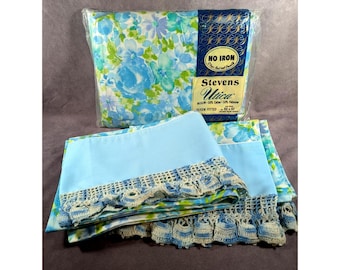 May include: A Stevens Utica queen fitted sheet in original plastic packaging with a blue and green floral pattern, labeled 60 x 80 inches. Below, a folded light blue sheet features a decorative cream and blue crocheted lace trim edge.