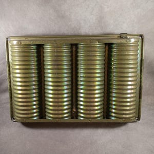 May include: A vintage metal container with four cylindrical, ribbed cans inside. The cans and the container have a metallic, olive-green hue. The container has a hinged lid and is rectangular in shape.