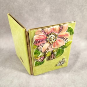 May include: A lime green, hinged compact with a raised floral design. The flower is pink with crystal and ruby accents, surrounded by green leaves. A small, jeweled butterfly is on the front.