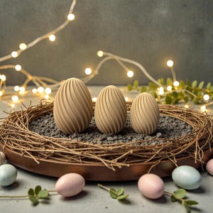 Könnte beinhalten: Drei beige, strukturierte Holzeier mit Spiralmuster liegen in einem Nest aus Zweigen und kleinen Steinen, umgeben von pastellfarbenen Eiern. Das Nest befindet sich in einer kupferfarbenen Schale. Lichterketten und Grünzeug im Hintergrund.