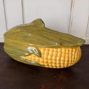 May include: A ceramic lidded serving dish shaped like a cob of corn. The base features bright yellow kernels, while the removable lid is molded to resemble green corn husks with a glossy finish and realistic textures throughout the entire piece.