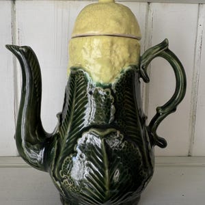 May include: A ceramic teapot featuring a dark green body with a textured, embossed leaf pattern and a contrasting pale yellow, crackle-glazed lid. The vessel includes a curved handle and a long, slender spout, showcasing a glossy finish throughout.