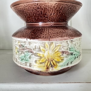 May include: A ceramic vase with a wide, flared rim and a rounded body. The upper section features a dark brown, textured wood-grain pattern. The lower section displays a cream-colored basket-weave relief with hand-painted yellow sunflowers and green leaves.