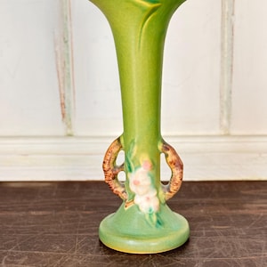 Pode incluir: Um vaso de cerâmica alto e esguio com um esmalte verde-lima vibrante e um acabamento craquelado visível. O vaso apresenta uma borda alargada e ondulada, e duas alças castanhas texturizadas em forma de ramo nas laterais, acentuadas com pequenos detalhes florais brancos e cor-de-rosa.