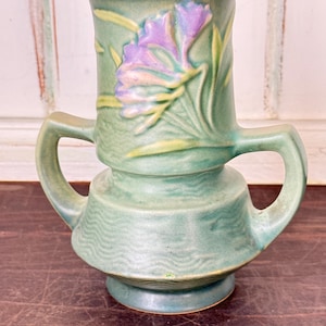 May include: A green ceramic vase featuring two side handles and a raised relief design of a purple flower with yellow leaves. The vase has a textured, wavy pattern around the lower body and a smooth, dark interior rim, standing on a circular base.
