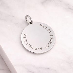 May include: A circular silver pendant featuring engraved geographic coordinates along the edge. The text reads 21°01'54.3"N and 89°08'17.18"W. The pendant includes a small jump ring at the top for attachment to a chain or cord.