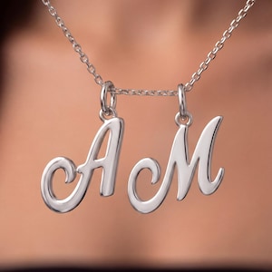 May include: A silver-toned necklace featuring two separate letter pendants, the capital letters A and M, rendered in a smooth, cursive script font. Each letter is suspended from the delicate silver-toned chain by a small, circular jump ring.