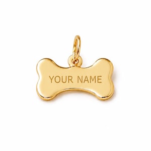 May include: A gold-colored, bone-shaped pet tag with the engraved text "YOUR NAME". The tag has a small loop at the top for attaching to a collar. The tag is made of a shiny, reflective material.