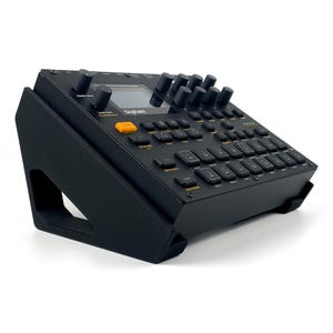 May include: A black Digitakt drum computer and sampler resting on a black angled stand. The device features a central screen, multiple rotary knobs, and a grid of backlit buttons with orange and white text labels, including a prominent orange button.
