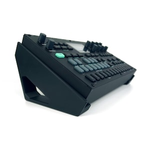 May include: A black electronic music synthesiser console resting on a matching angled black stand. The device features a grid of rectangular buttons, several circular control knobs, and a single prominent light green square button on the top surface.