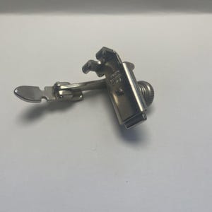May include: A silver sewing machine presser foot. The presser foot has a rectangular body with a rounded knob and a hinged arm with a small, flat, oval-shaped foot. The presser foot is designed to hold fabric in place while sewing.