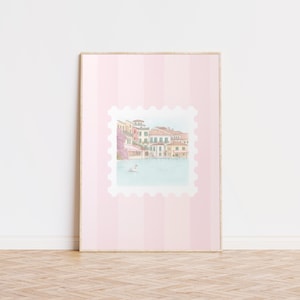 Pu&ograve; includere: Una stampa incorniciata con un'illustrazione ad acquerello di un villaggio sul lago con edifici colorati e un cigno. La stampa &egrave; ambientata su uno sfondo a righe rosa, che ricorda un grande francobollo.