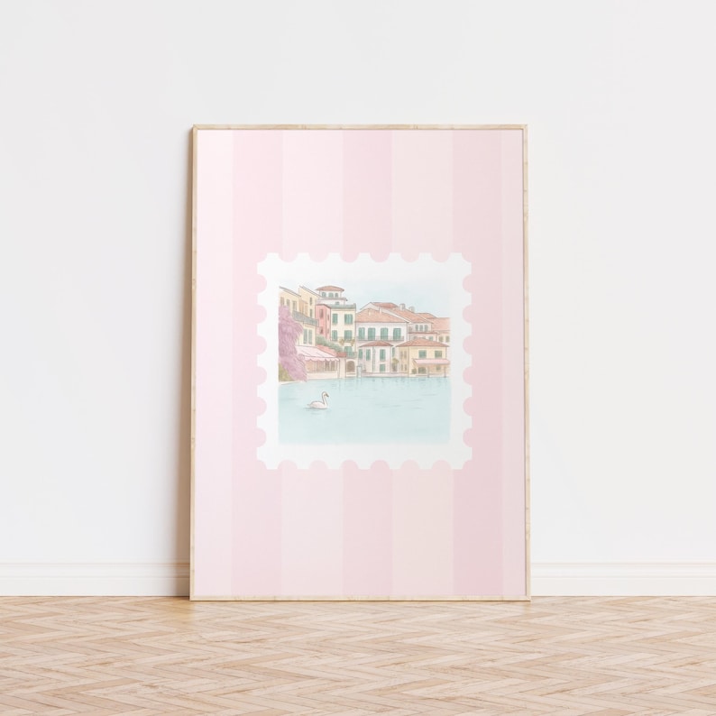 Pu&ograve; includere: Una stampa incorniciata con un'illustrazione ad acquerello di un villaggio sul lago con edifici colorati e un cigno. La stampa &egrave; ambientata su uno sfondo a righe rosa, che ricorda un grande francobollo.