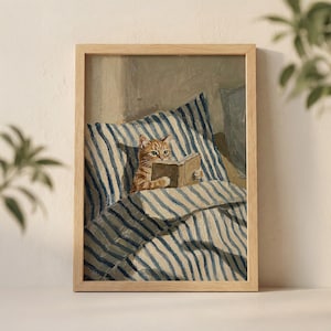 Könnte beinhalten: Ein gerahmter Ölgemäldedruck, der eine orangefarbene getigerte Katze zeigt, die im Bett unter einer blau-weiß gestreiften Decke und einem Kissen liegt und ein offenes Buch hält. Das Kunstwerk befindet sich in einem hellen Holzrahmen.