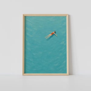 May include: A framed art print featuring a painting of a figure in a red swimsuit floating in calm, turquoise water. The artwork is enclosed in a light-colored wooden frame, capturing a top-down view of the swimmer surrounded by ripples.