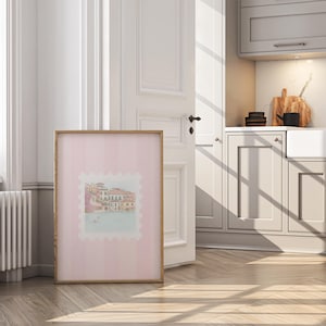 Pu&ograve; includere: Una stampa incorniciata con un'illustrazione ad acquerello di un paesaggio urbano europeo, su uno sfondo rosa a righe. L'opera d'arte &egrave; in una cornice di legno chiaro e si appoggia a una porta e una parete bianche, con uno scorcio di una cucina sullo sfondo.