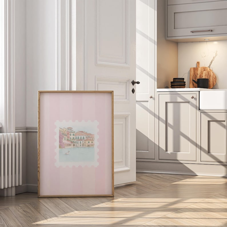 Pu&ograve; includere: Una stampa incorniciata con un'illustrazione ad acquerello di un paesaggio urbano europeo, su uno sfondo rosa a righe. L'opera d'arte &egrave; in una cornice di legno chiaro e si appoggia a una porta e una parete bianche, con uno scorcio di una cucina sullo sfondo.