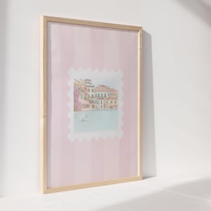 Pu&ograve; includere: Stampa artistica incorniciata con un'illustrazione ad acquerello di un paesaggio europeo sul lungomare. L'opera d'arte &egrave; ambientata su uno sfondo a righe rosa e incorniciata in legno naturale. Un cigno galleggia sull'acqua.