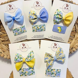 May include: Four sets of hair bows with matching clips. The bows are made of various fabrics, including floral print, gingham, and solid colors. The clips are all rectangular and have a floral print. The text on the cards reads "Yellow Belles Boutique" and includes social media information.